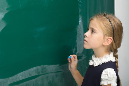 Adorable schoolgirl writing on green chalkboard. Side view of elementary school student girl standing next to blackboard in classroom. School and education concept