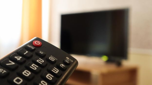 Hand holding a television remote control and surfing programs on television. watch, turn on or off the TV in the living room or bedroom on the black-screen nightstand. Copy space