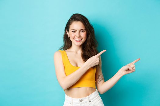 Summer lifestyle concept. Cheerful stylish woman showing direction, pointing fingers right, that way gesture, standing over blue background.