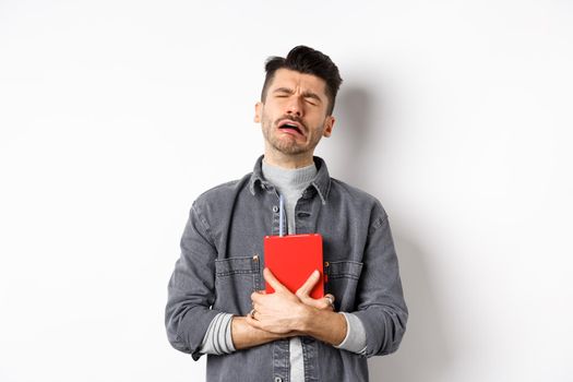 Sad cryig man holding red diary and sobbing, miserable guy carry journal with him, standing against white background.