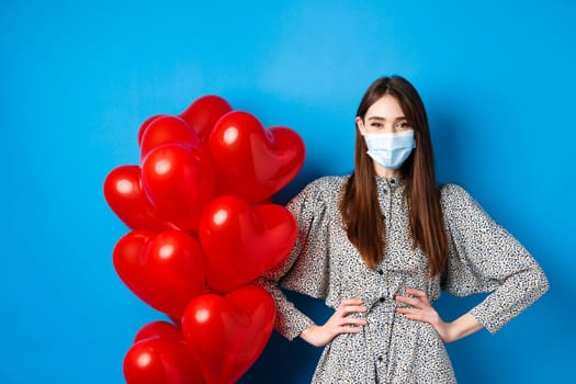 Pandemic and Valentines day. Cheerful smiling girl in medical mask, standing near romantic heart balloons and looking at camera, wearing dress, blue background.