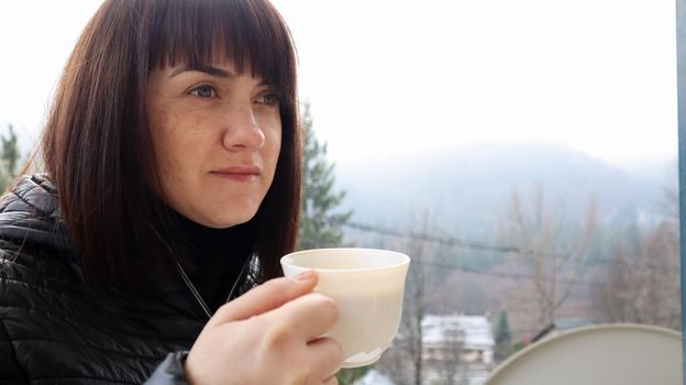 Beautiful happy woman with a white cup of coffee or tea enjoying the beautiful view of the mountain landscape in the morning fog from the balcony of her hotel room. Autumn and winter travel concept.
