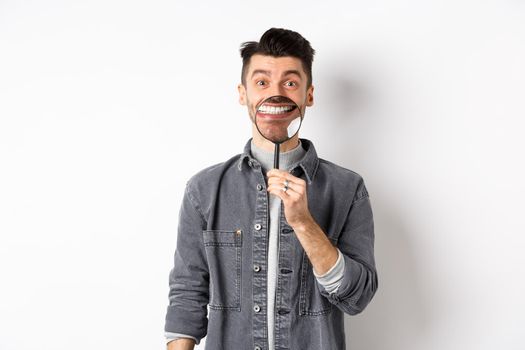 Handsome positive guy showing white perfect smile with magnifying glass, standing against white background.