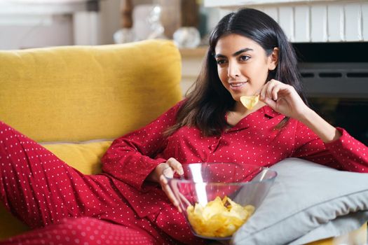 Persian woman at home watching TV. Girl eat chips potatoes