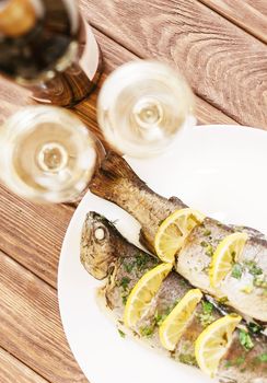 Fish dish on a plate on wooden table and glasses of white wine for couple, top view.