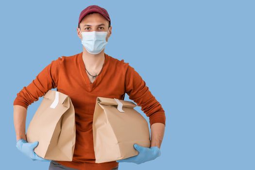 Young delivery man in medical mask holding and carrying a cardbox isolated on blue background. Buy food online in quarantine concept