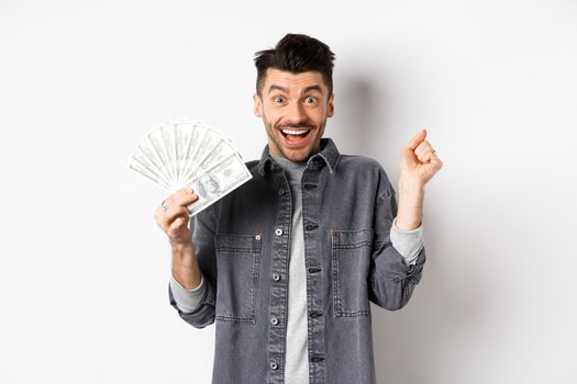 Cheerful young man jumping from excitement and showing dollar bills, winning prize cash, making money and rejoicing, standing on white background.