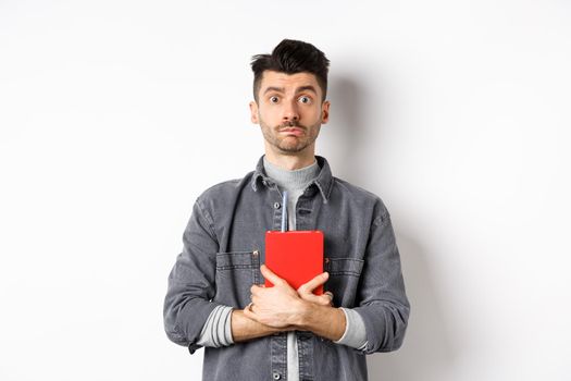 Cute and shy guy hugging his diary, holding red journal and look modest at camera, standing against white background.