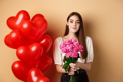 Beautiful girl holding bouquet of pink roses and smiling at camera, going on romantic date, standing near valentines heart balloons, beige background.