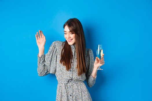 Celebration and party concept. Dancing girl holding glass of champagne, having fun at holiday, celebrating birthday, standing on blue background.