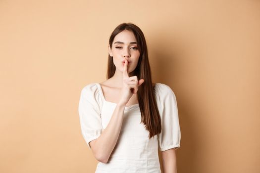 Sensual cute girl hiding secret, showing hush gesture, make shhh sound with finger on lips, stay quiet sign, standing on beige background.
