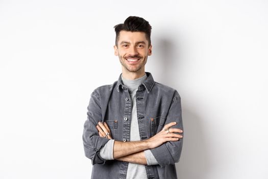 Handsome young man smiling with arms crossed on chest, feeling ready, looking motivated at camera, standing on white background.