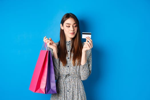 Beautiful girl going on shopping with plastic credit card, holding paper bags with purchased items, standing pleased on blue background.