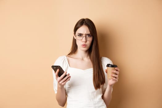 Confused girl in glasses frowning at camera after reading mobile phone, holding coffee cup, standing displeased on beige background.