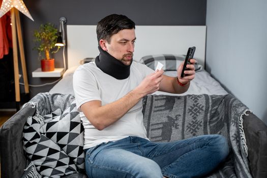 Patient with collar and neck injury receives an online video consultation with doctor and holds pill in hand. Man in foam cervical collar calls traumatologist on video and shows medications on phone.
