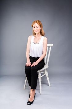 beautiful young caucasian woman with long red hair in high heels sitting on a chair, black trousers and white shirt in full growth on a gray background. Business concept.
