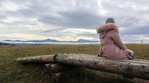 A woman sits with her back on wooden logs high in the mountains in cold autumn weather and enjoys the beautiful view of the mountainous natural landscape