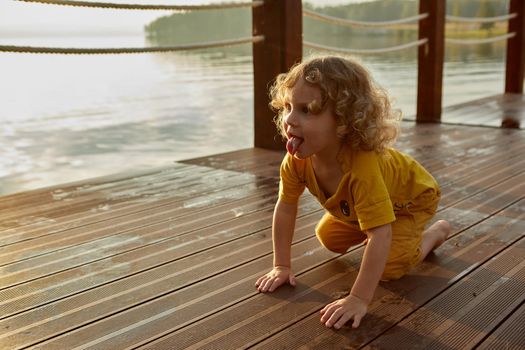 Adorable toddler with curly hair showing tongue and crawling on wet quay near lake water at sunset