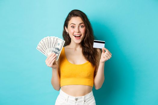 Attractive fit woman in summer outfit, showing dollar bills money and plastic credit card, smiling amazed, standing against blue background.