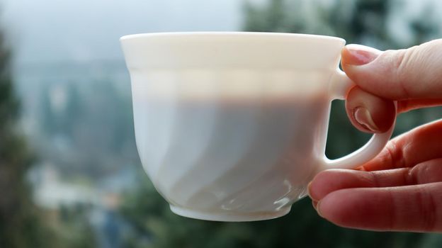 Girl hand holds a cup of white coffee and has a blurred green nature background. Mug in a female hand on a background of mountains