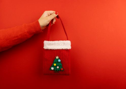 Caucasian woman hand at red winter sweater holding red knitted Christmas bag at red background, creative Christmas shopping and new year presents concept, copy space.
