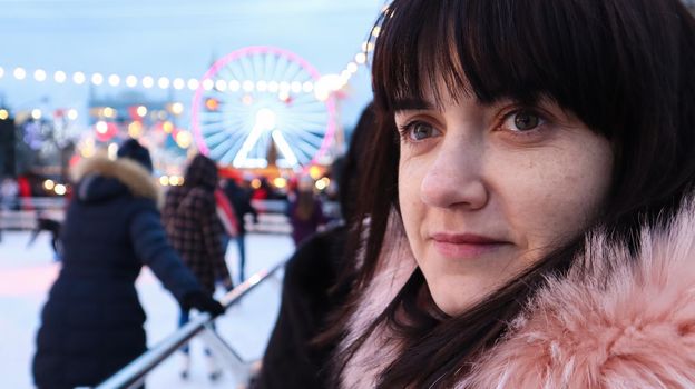 Portrait of a happy young woman in winter on a skating rink in a pink jacket with fur outdoors during the day. Entertainment at the New Year's fair. Woman looking at ice rink, Christmas holiday
