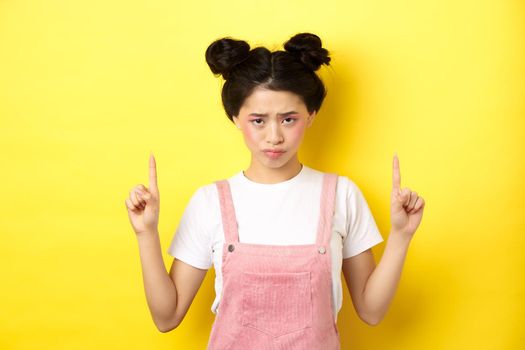 Sad and gloomy asian woman frowning, pointing fingers up and complaining on unfair thing, standing upset on yellow background.