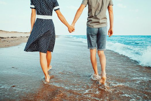 Young loving couple walking on shore on sea waves and holding hands, beach summer vacations.
