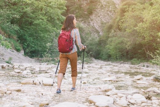 Hiker backpacker young woman with trekking poles walking along the river gorge in summer.