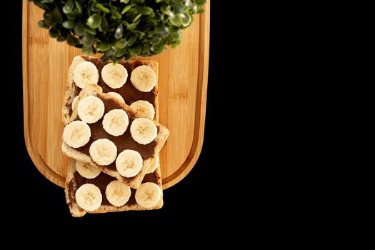 Three banana white bread toasts spread with chocolate butter that lie on a chopping board with a sprig of leaves on a dark background. top view with area for text