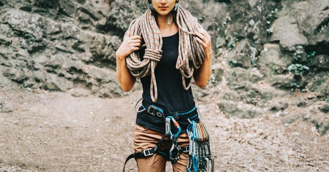 Sporty woman wearing in safety harness with climbing equipment holding rope on background of rock.