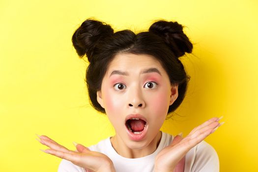 Close-up of excited asian woman with glamour makeup and hairstyle, screaming of amazement, open mouth and look at camera in awe, yellow background.