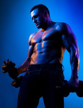 Muscular bodybuilder guy doing exercises with dumbbells isolated on blue neon. Naked athlete with strong body. Shirtless hunk with perfect abs