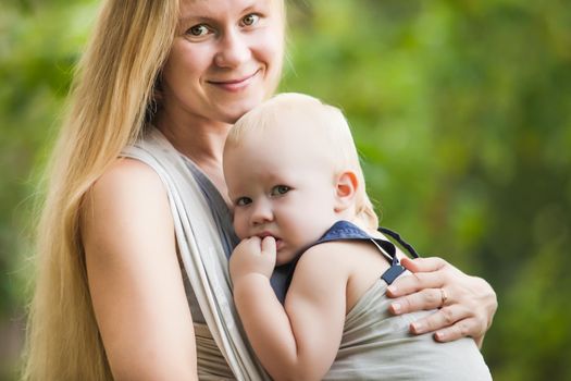 Mother is carrying her child and walking. Baby in sling outdoor.