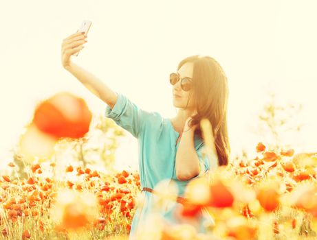 Beautiful young woman in sunglasses taking selfie with smartphone in poppies meadow on sunny summer day.