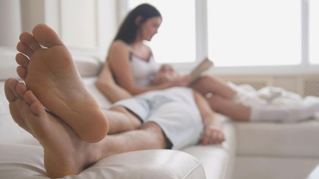 Happy young attractive couple are together on the couch, the girl reads a book, the guy lies on her lap, people's attitudes