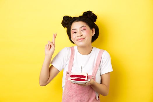 Holidays and celebration. Positive asian birthday girl cross fingers, making wish with b-day cake and lit candle, smiling happy at camera, yellow background.