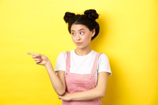 Portrait of stylish asian woman with makeup and summer clothes, pointing and looking left at logo, standing intrigued on yellow background.