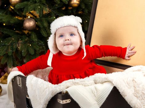 The baby is sitting in an old suitcase. A child in a red Christmas dress and a white hat sits in a suitcase and expresses emotions against the background of garlands and a Christmas tree
