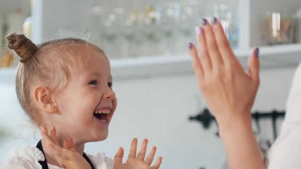 Happy little girl with her mother clap their hands in the kitchen, close up