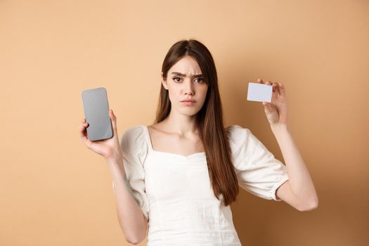 Sad and disappointed girl showing empty smartphone screen and plastic credit card, complaining and frowning, standing on beige background.