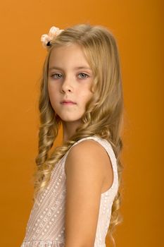 Beautiful blonde girl looking at camera. Adorable seven years old girl with serious face expression. Portrait of cute gray-eyed child wearing nice dress dress posing half sided on ochre background