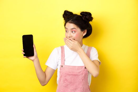 Online shopping concept. Silly japanese girl with beauty makeup, cover mouth with hand laughing and showing empty smartphone screen, show funny thing on phone, yellow background.