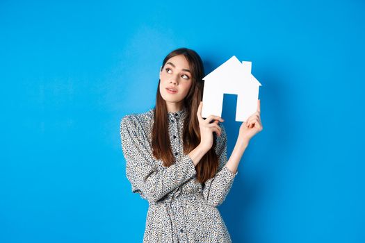 Real estate, realtors and insurance concept. Dreamy beautiful adult woman thinking of own house, showing paper home cutout and look aside at logo, standing against blue background.