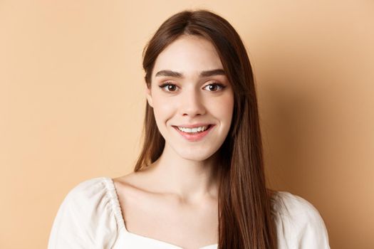 Close up portrait of happy woman with long hair and natural makeup, smiling cheerful at camera, standing on beige background.