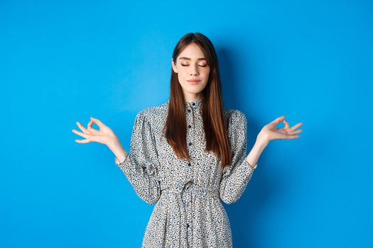 Mindful young woman in dress, close eyes and smile, meditating peaceful, standing with hands spread sideways with zen sign, blue background.