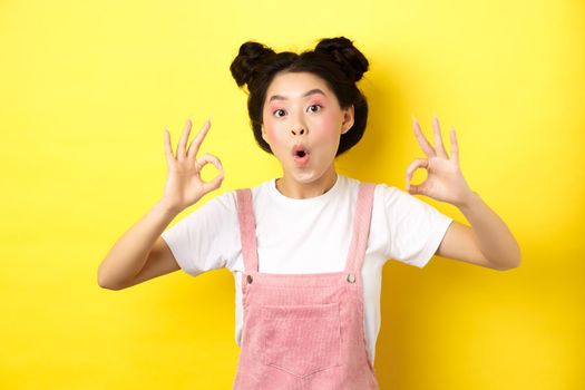 Excited beauty girl with bright makeup and summer clothes, showing okay signs and say wow, praise good promo deal, approve advertisement, yellow background.