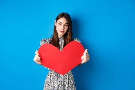 Valentines day and relationship concept. Tender young woman in dress stretch out hand and giving big red heart to you, making confession, standing romantic on blue background.