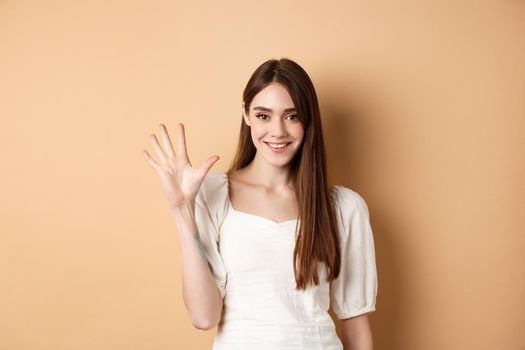 Attractive young woman show fingers number five, smiling and looking confident, standing on beige background.