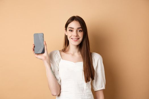 Happy girl showing empty mobile screen and smiling, demonstrate phone app, standing on beige background.